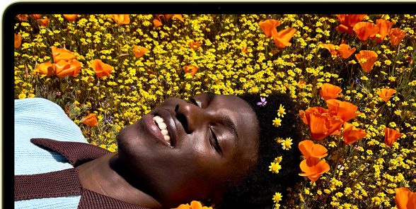 MacBook Neo, Farbe Zitrus, das Display zeigt eine Person, die in einer Blumenwiese in der Sonne liegt und lächelt
